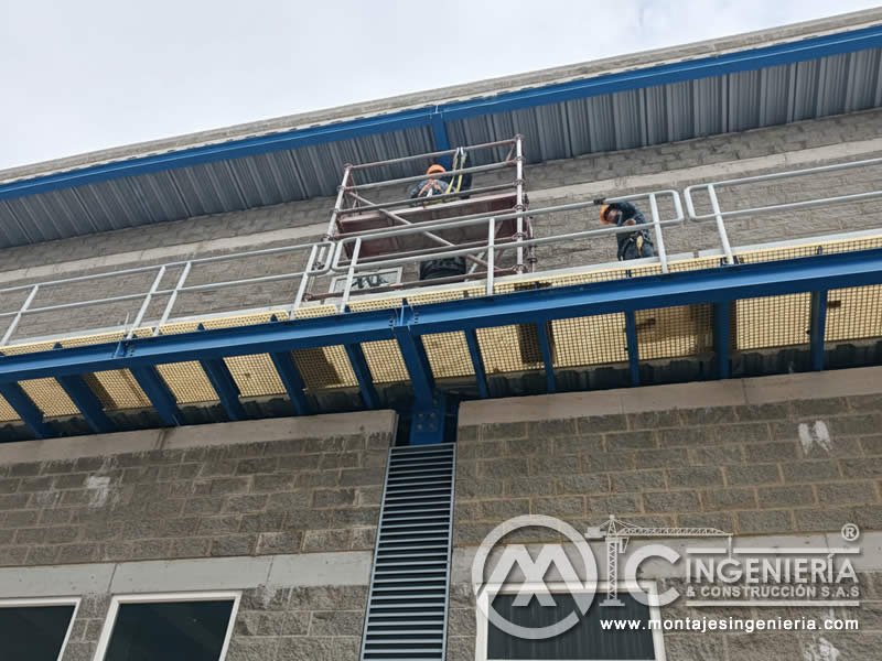 Dos técnicos en plataforma FRP azul finalizando montaje en fachada industrial en Bogotá, Colombia. Montajes, Ingeniería y Construcción MIC SAS.